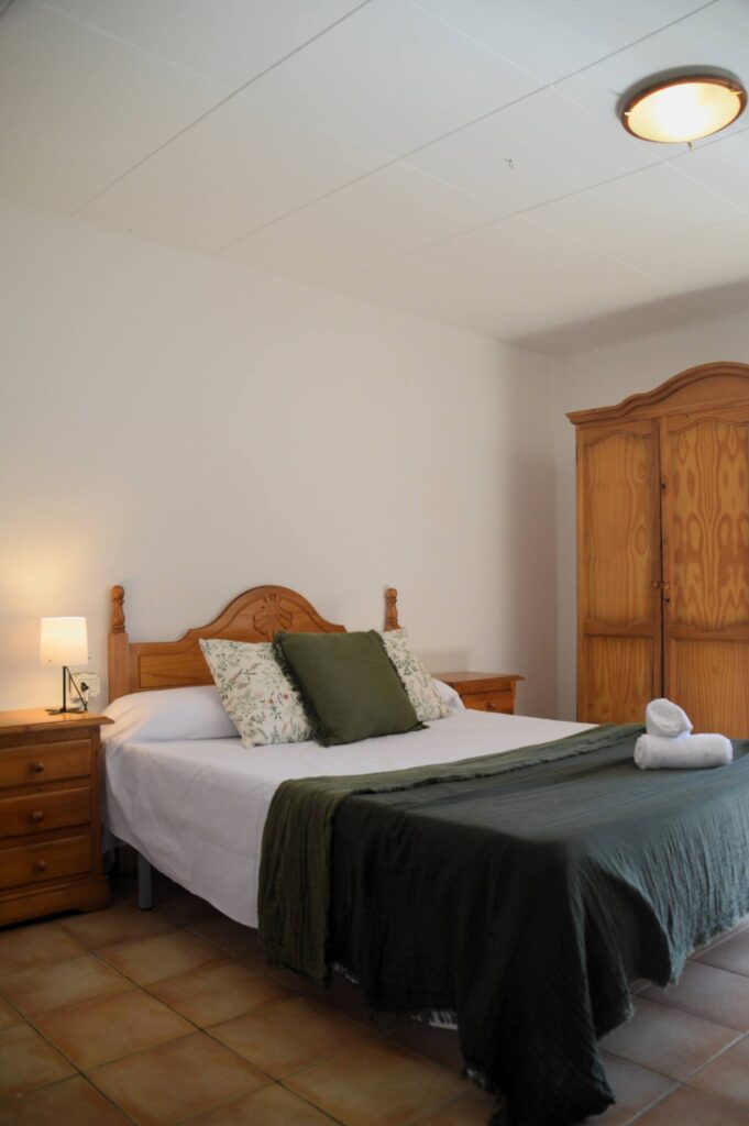 Una altra vista del dormitori que mostra el llit doble amb cobrellit verd i coixins a joc, una tauleta amb llum i un armari de fusta, reflectint l’ambient rústic de la casa