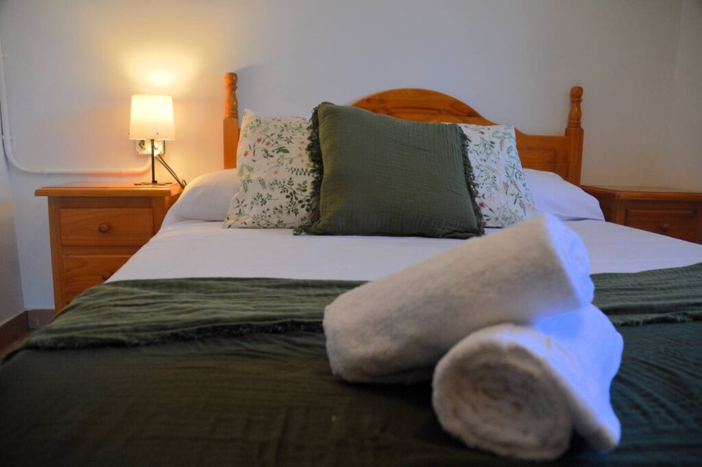 Una altra vista del dormitori que mostra el llit doble amb cobrellit verd i coixins a joc, una tauleta amb llum i un armari de fusta, reflectint l’ambient rústic de la casa