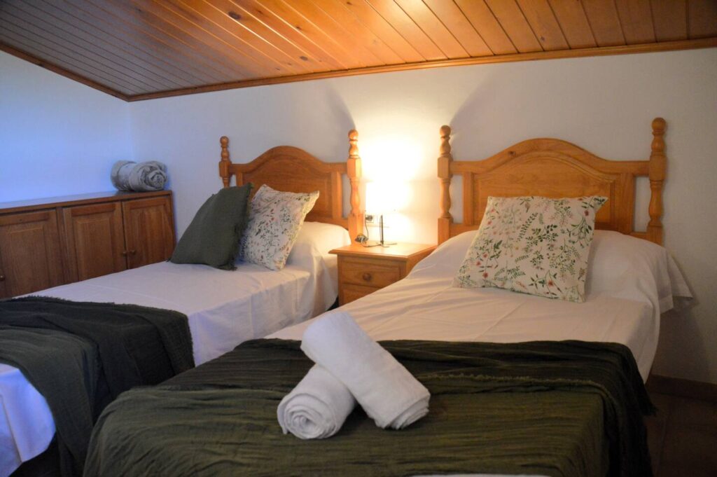 Una altra vista del dormitori que mostra el llit doble amb cobrellit verd i coixins a joc, una tauleta amb llum i un armari de fusta, reflectint l’ambient rústic de la casa