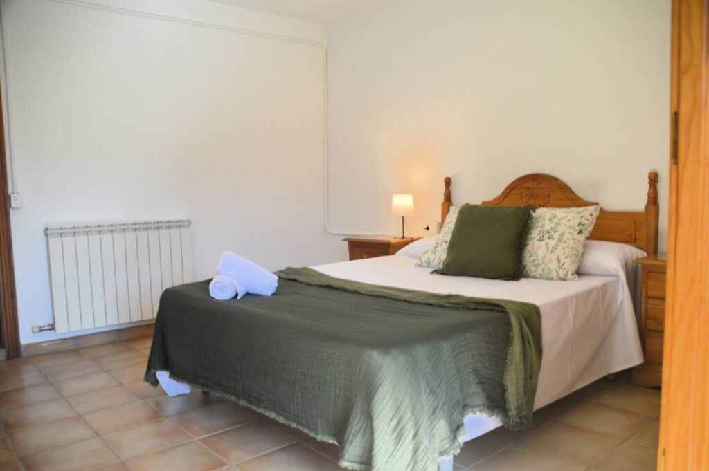 Una altra vista del dormitori que mostra el llit doble amb cobrellit verd i coixins a joc, una tauleta amb llum i un armari de fusta, reflectint l’ambient rústic de la casa