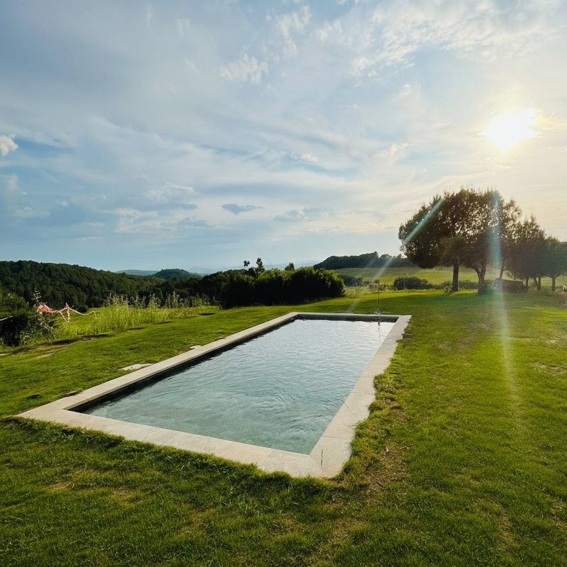 Piscina rectangular exterior envoltada de gespa amb camps i arbres al capvespre.