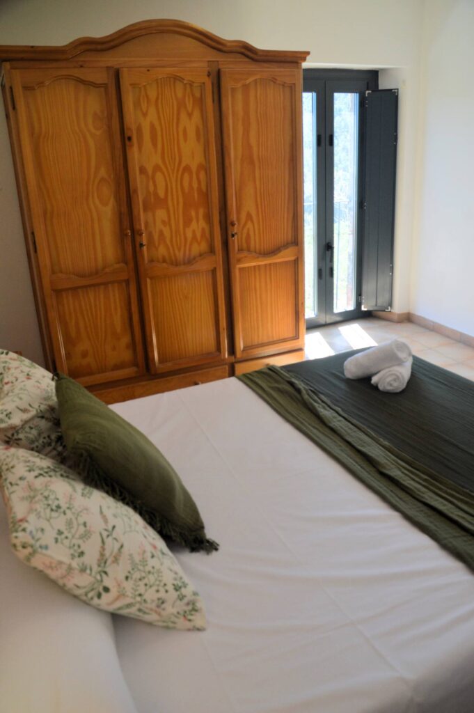 Una altra vista del dormitori que mostra el llit doble amb cobrellit verd i coixins a joc, una tauleta amb llum i un armari de fusta, reflectint l’ambient rústic de la casa