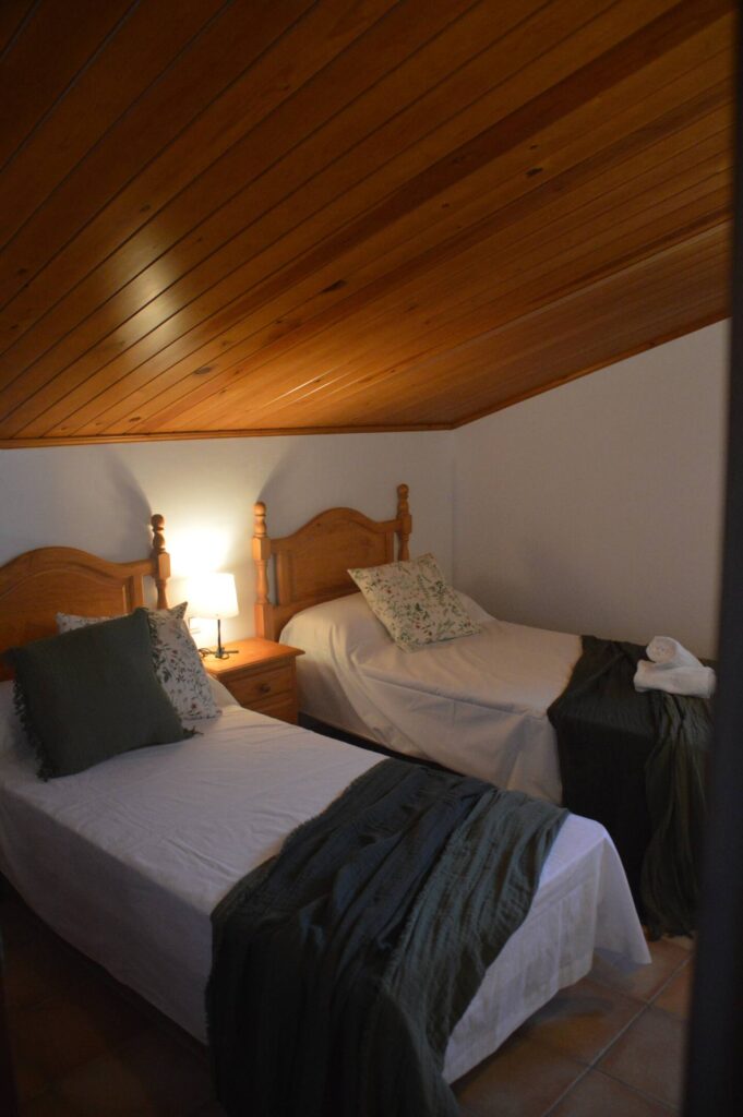 Una altra vista del dormitori que mostra el llit doble amb cobrellit verd i coixins a joc, una tauleta amb llum i un armari de fusta, reflectint l’ambient rústic de la casa