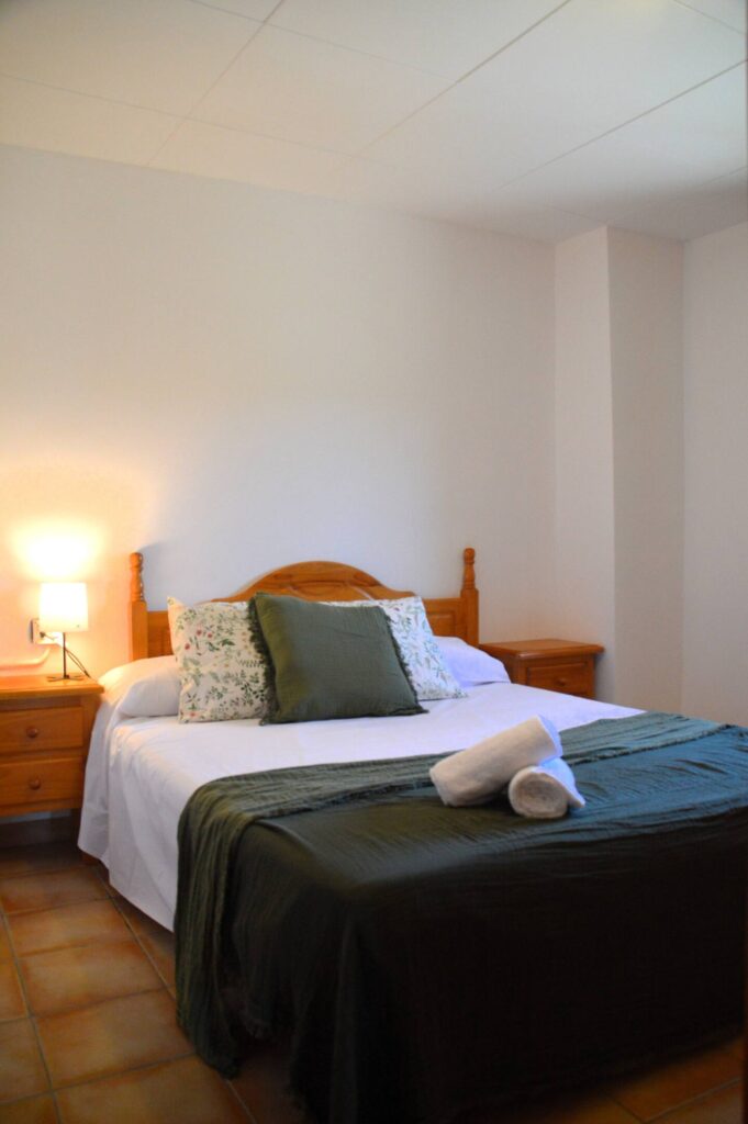 Una altra vista del dormitori que mostra el llit doble amb cobrellit verd i coixins a joc, una tauleta amb llum i un armari de fusta, reflectint l’ambient rústic de la casa